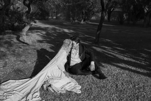Wedding couple portrait in black and white, bride and groom sitting on grass sharing a forehead kiss under tree shade, lace veil and long train