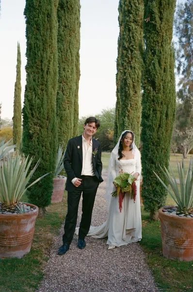 Couple portrait of bride and groom on a gravel garden path, bride holding bouquet with veil flowing, cypress trees behind