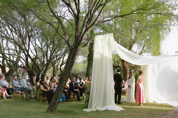 Outdoor wedding ceremony with bride and groom at a white draped altar, guests in wooden chairs beneath trees on a lawn, daylight sky