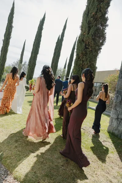 Wedding processional with bridesmaids walking down aisle on a grassy lawn, carrying bouquets as bride in long veil follows under cloudy sky