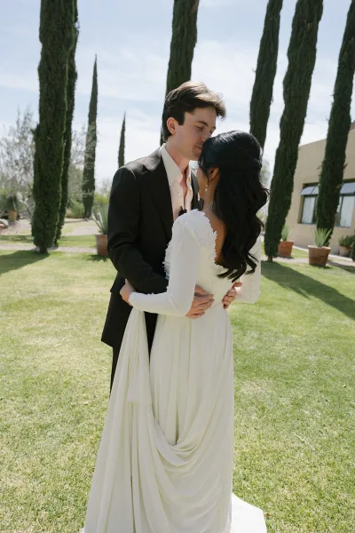 Wedding couple portrait of bride and groom embracing with foreheads touching, bride in long sleeve lace bodice gown on lawn by cypress trees under blue sky