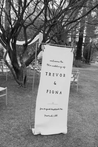Wedding welcome sign with calligraphy lettering on a fabric banner hanging from a metal stand at an outdoor garden ceremony entrance