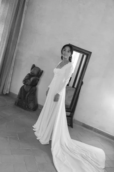 Bridal portrait in black and white of a bride in a long sleeve lace bodice gown with train, posed by a leaning mirror and stucco doorway