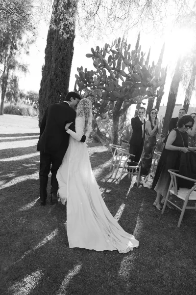 Ceremony recessional as newlyweds walk down the aisle, bride’s lace veil and long train flowing past guest chairs on a sunny lawn with trees and cactus