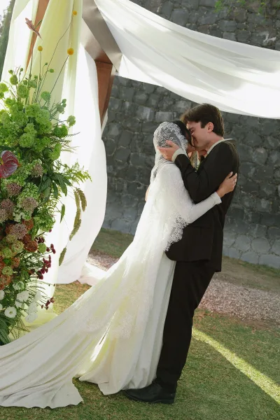 Wedding kiss with lace veil accent as the bride and groom embrace beneath a draped arch with florals and greenery by a stone wall lawn