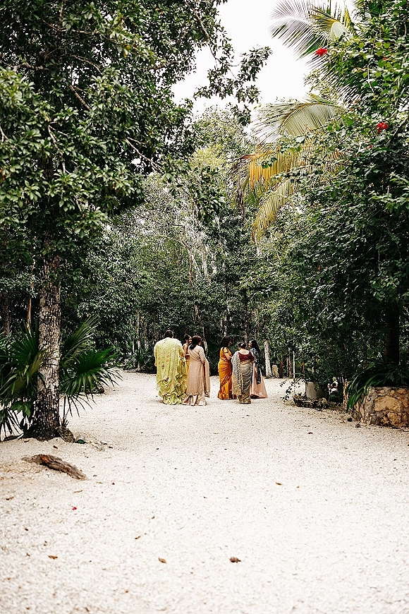 Wedding guests in colorful sarees walk down a white gravel path under tropical palm fronds, one wearing an embroidered shawl