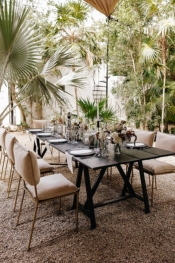 Reception tablescape with an outdoor reception table on black farm tables, bud vase florals and candles under string lights by palms