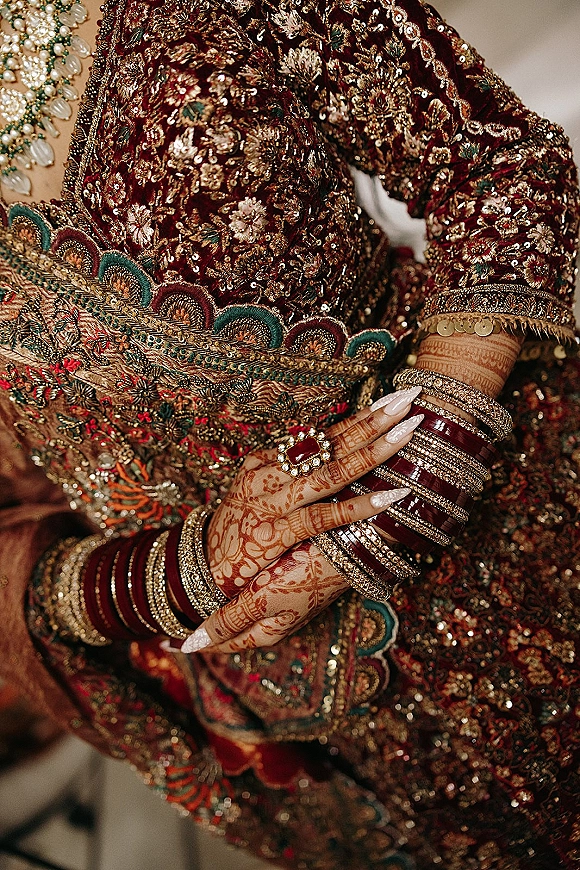 Bridal outfit details close up of mehndi hands showing red lehenga gold embroidery, dupatta border sequins, bangles, pearl ring in soft indoor light