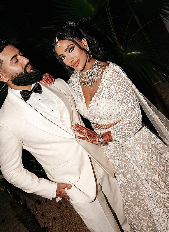Couple portrait of bride leaning on groom in a white tuxedo with black bow tie, henna hands and veil under palm leaves at night