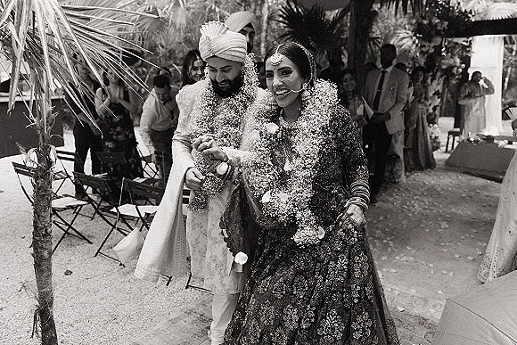 Wedding recessional as bride and groom walking hand in hand, floral garlands on, rose petals falling on a garden aisle with guests seated