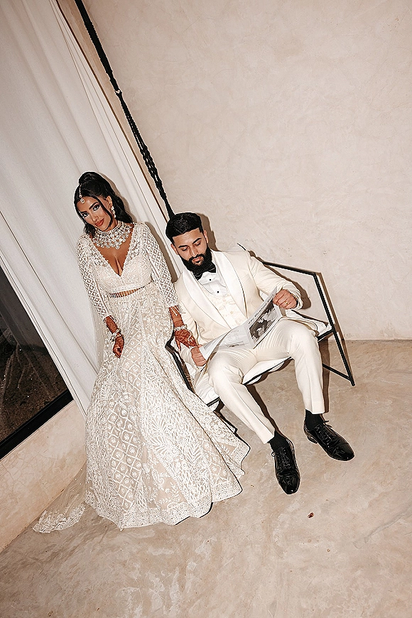 Couple portrait with bride in white embroidered gown and henna, standing beside groom in white tuxedo reading a newspaper on a chair by a staircase