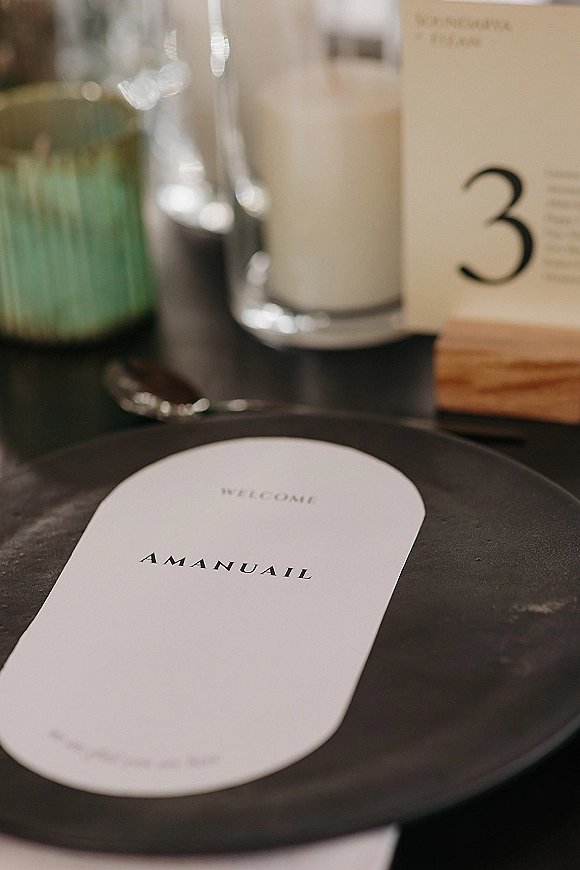 Wedding table number card in a wooden holder beside a black plate, glassware, cutlery, and candle on a reception tablescape