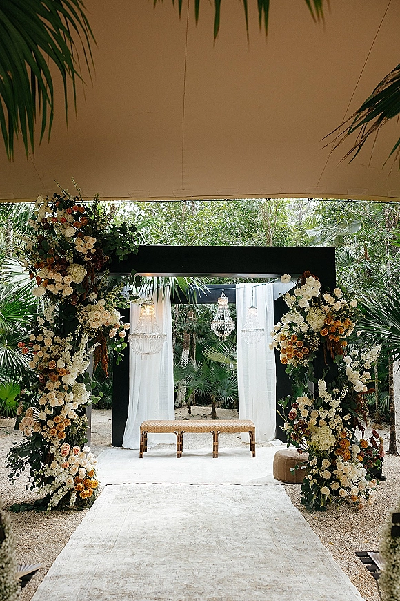 Wedding ceremony backdrop with a modern wedding altar featuring a black square arch, draped fabric, chandeliers, and floral greenery in a palm garden