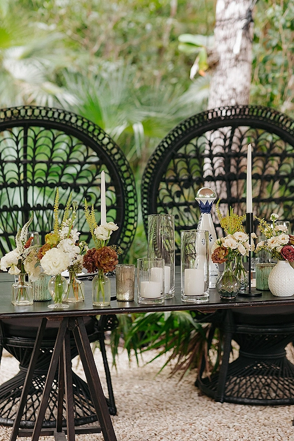 Reception tablescape with bud vase wedding centerpiece, black taper candles, mixed glass and ceramic vases on a dark table amid palms