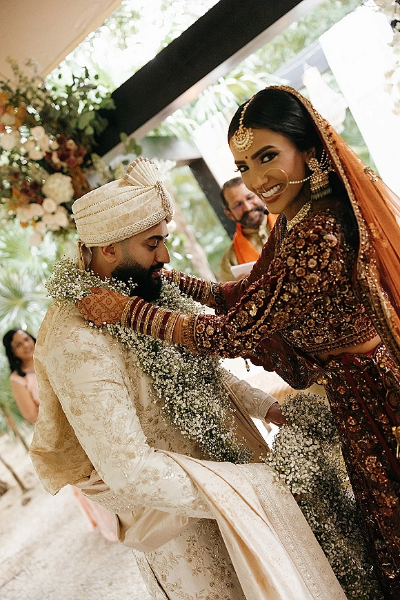 Ceremony moment at an Indian wedding ceremony as the bride in a maroon lehenga places a floral garland on the groom under an indoor floral arch