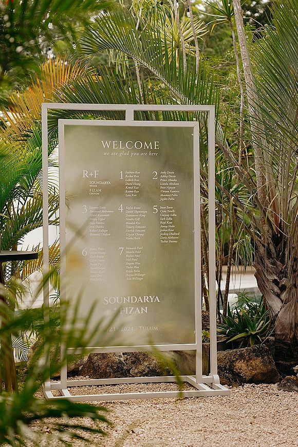 Wedding seating chart on a clear acrylic sign in a white metal frame, set on gravel with palm trees and tropical foliage behind