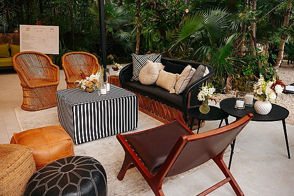 Wedding lounge area with wedding lounge seating, wicker chairs and black sofa around a striped ottoman, candles and florals amid palms on a gravel patio