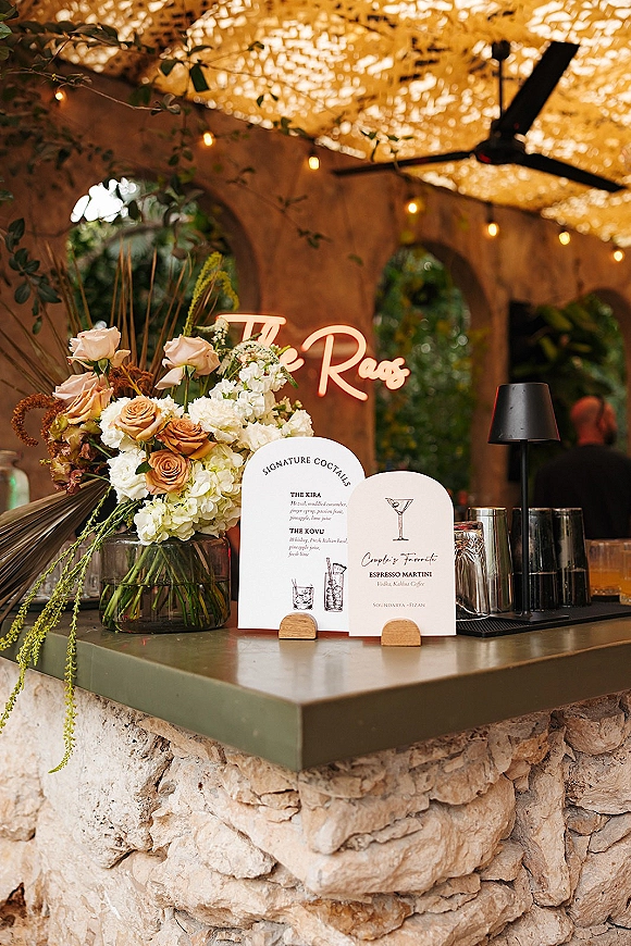 Wedding bar signage and signature cocktail menu cards on a stone outdoor bar with roses and hydrangeas, neon sign, and string lights under stucco arches