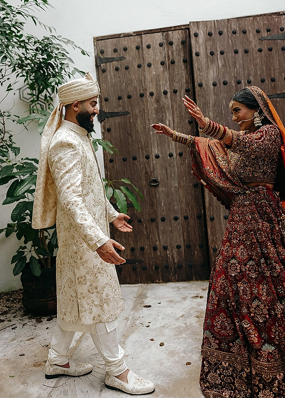 Wedding first look as South Asian bride in red lehenga and gold jewelry reaches for groom in sherwani by wooden doors and potted plant