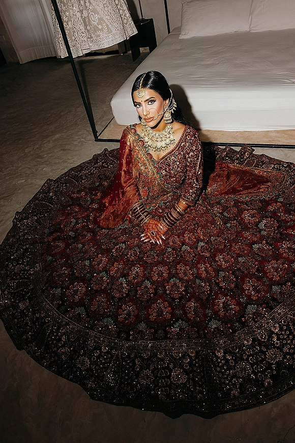 Bridal portrait of a South Asian bride in a red embroidered lehenga and gold jewelry, sitting on a bedroom floor by a bed