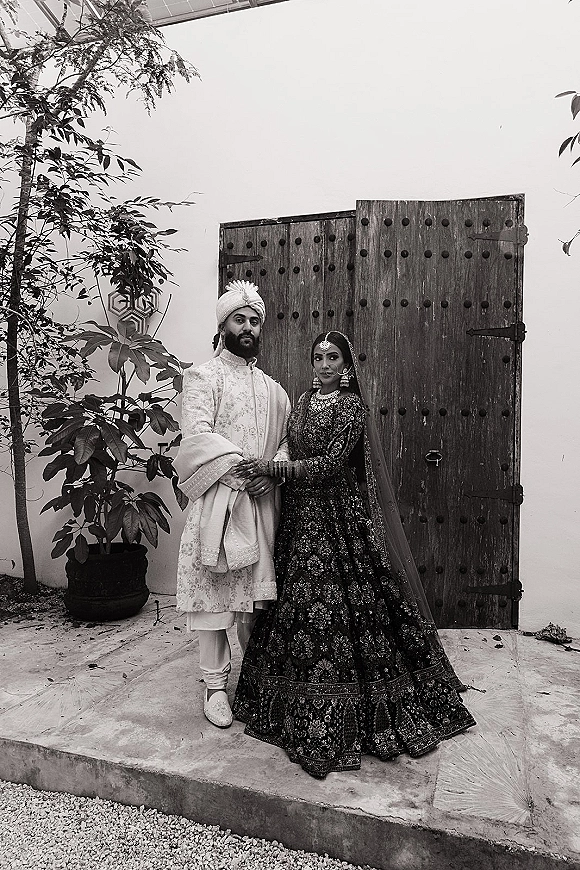 Wedding couple portrait of a south asian wedding couple holding hands, bride in embroidered lehenga and dupatta veil by wooden doors in a courtyard