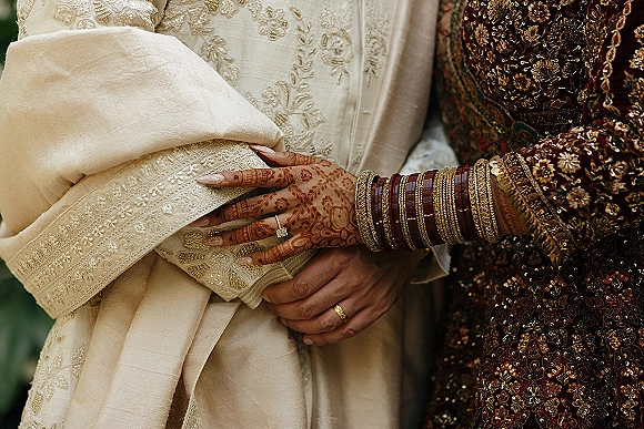 Wedding couple close-up as bride holding groom arm, showing henna hands with bangles and rings against a softly blurred background