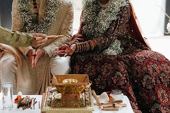 Wedding ceremony ritual as bride and groom in garlands and embroidered lehenga and sherwani perform rites with ritual bowl on a white table indoors