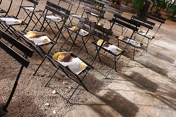 Ceremony seating with outdoor ceremony chairs lined in rows, paper fan accents, and rose petal aisle runner on gravel under palm trees