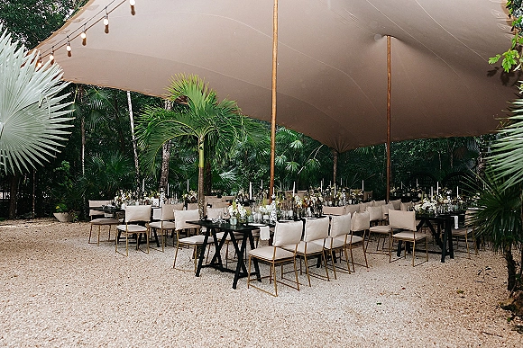 Reception tablescape at an outdoor tented reception with long banquet tables, taper candles, floral centerpieces, and string lights under palm trees