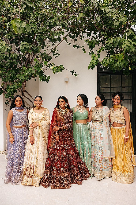 Bridesmaids portrait of women in colorful lehenga outfits with gold jewelry and dupattas posed by a white wall and window in a courtyard
