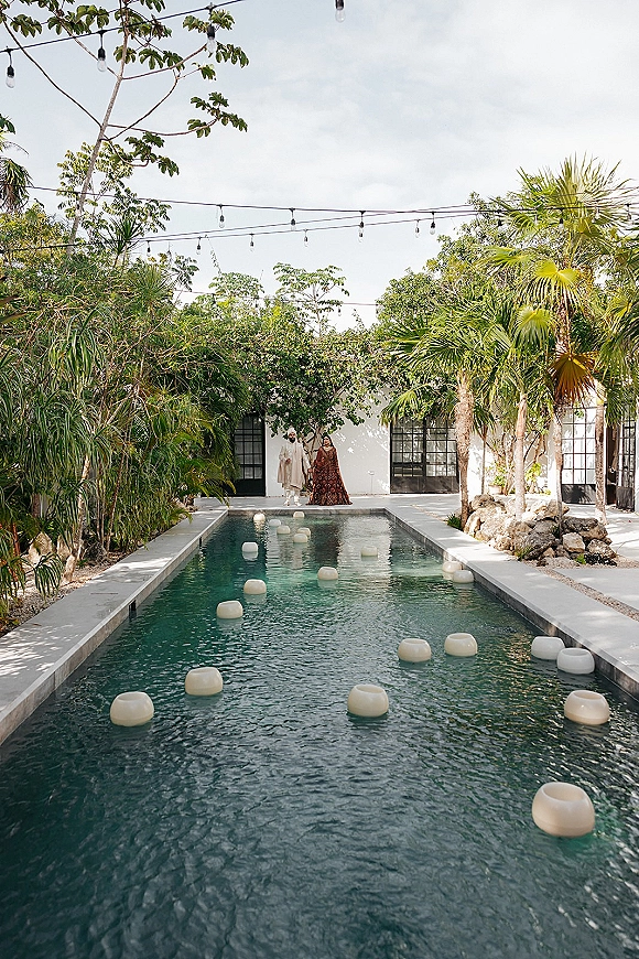 Couple portrait of bride and groom by pool in wedding attire, framed by string lights and floating candles in a tropical villa courtyard