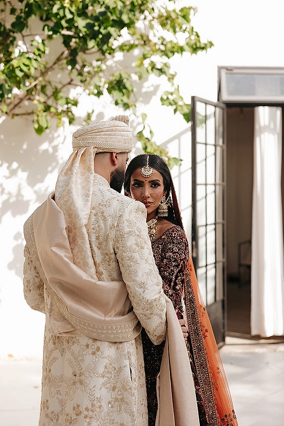 Couple portrait of a South Asian wedding couple embracing, groom in turban and sherwani, bride in maroon lehenga by sunlit white wall patio