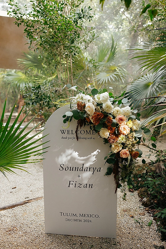 Wedding welcome sign with an arched board and roses, set on a gravel path in a tropical garden with eucalyptus greenery