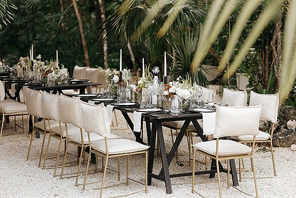 Reception tablescape with an outdoor wedding reception table on black farm tables, lined with taper candles and floral centerpieces amid palm trees