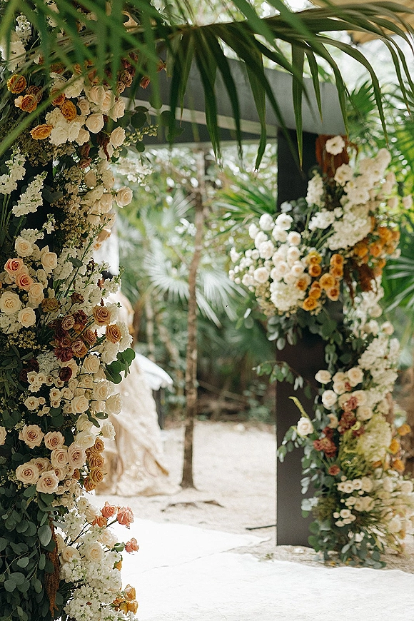 Wedding ceremony arch with asymmetrical roses, white and yellow flowers, and eucalyptus on a modern black frame amid palm trees and sand