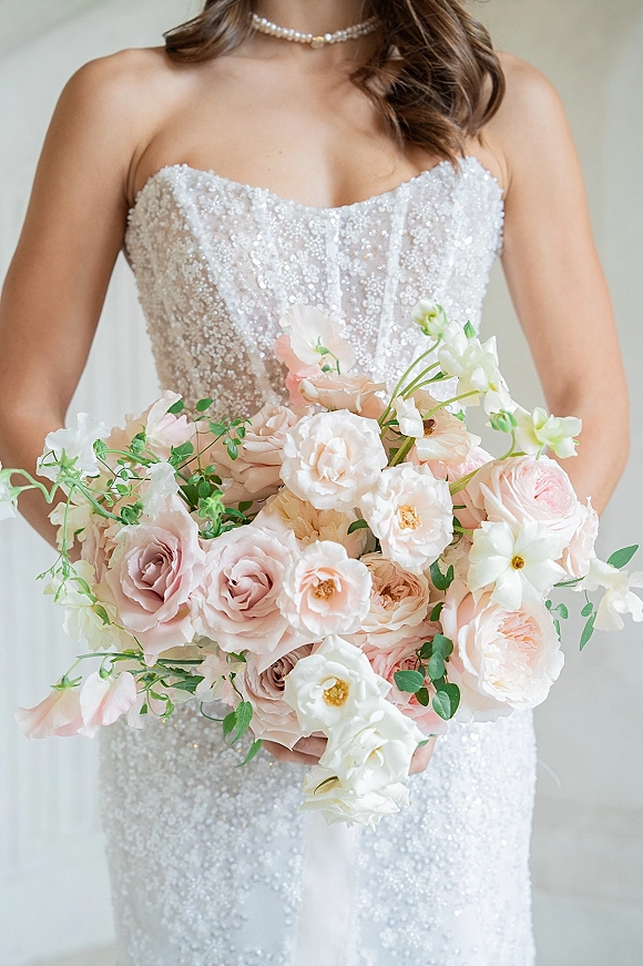 Bridal bouquet of blush and white roses with sweet pea and greenery held against a strapless beaded wedding dress and pearl choker on neutral wall