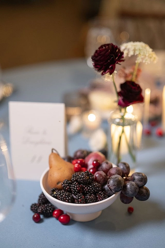 Wedding fruit display with a fruit bowl of grapes, blackberries, cranberries and pear beside a burgundy bud vase on a candlelit reception table
