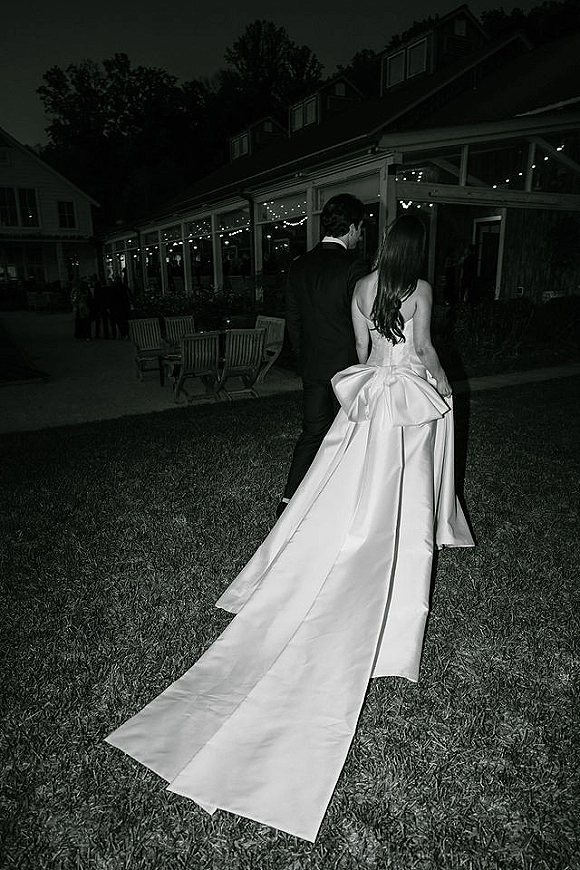 Couple portrait of bride and groom walking away, her long train and bow visible, under string lights on an outdoor patio at night