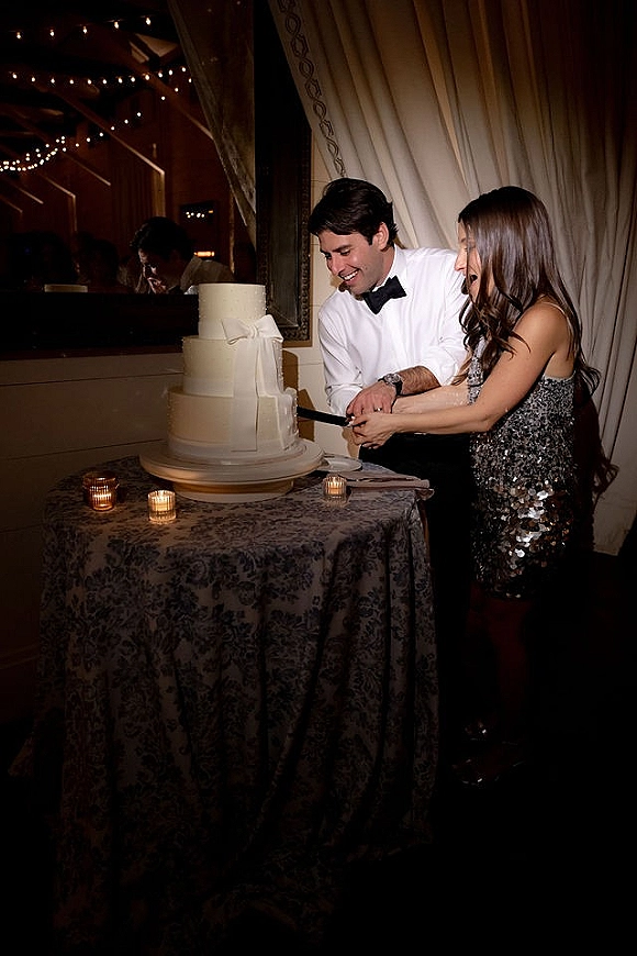 Wedding cake cutting as bride in sequin dress and groom in bow tie slice a tiered cake with ribbon on a candlelit table under string lights