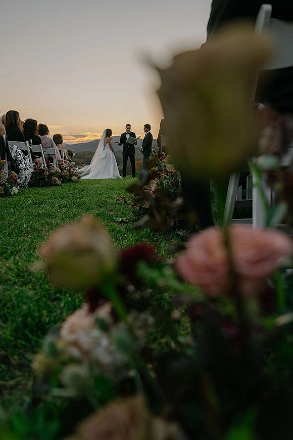 Wedding ceremony with bride and groom at a floral arch as officiant leads vows, long veil trailing along aisle flowers at sunset lawn with hills