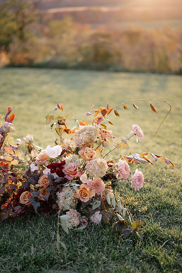 Wedding bouquet with garden rose blooms and trailing greenery, resting on grass in warm sunset light with trees and hills behind