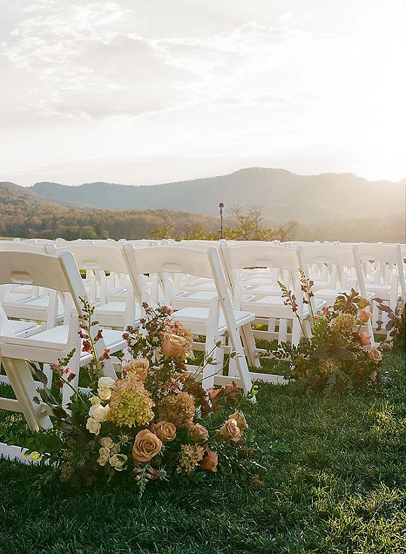 Ceremony aisle decor with roses, hydrangea and greenery lining white folding chairs on a grassy lawn with mountain view at sunset