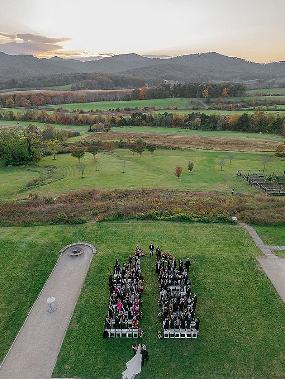 Outdoor wedding ceremony with white ceremony chairs lining an aisle, guests gathered as bride walks toward groom in a grassy field at sunset