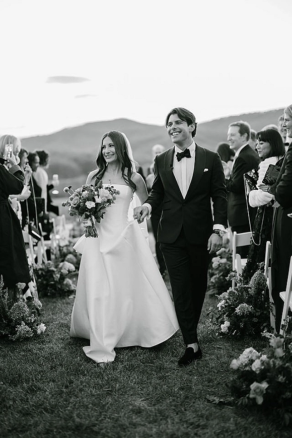 Wedding recessional as newlyweds walking down aisle holding hands, bride with bouquet, guests cheering on a mountain lawn setting