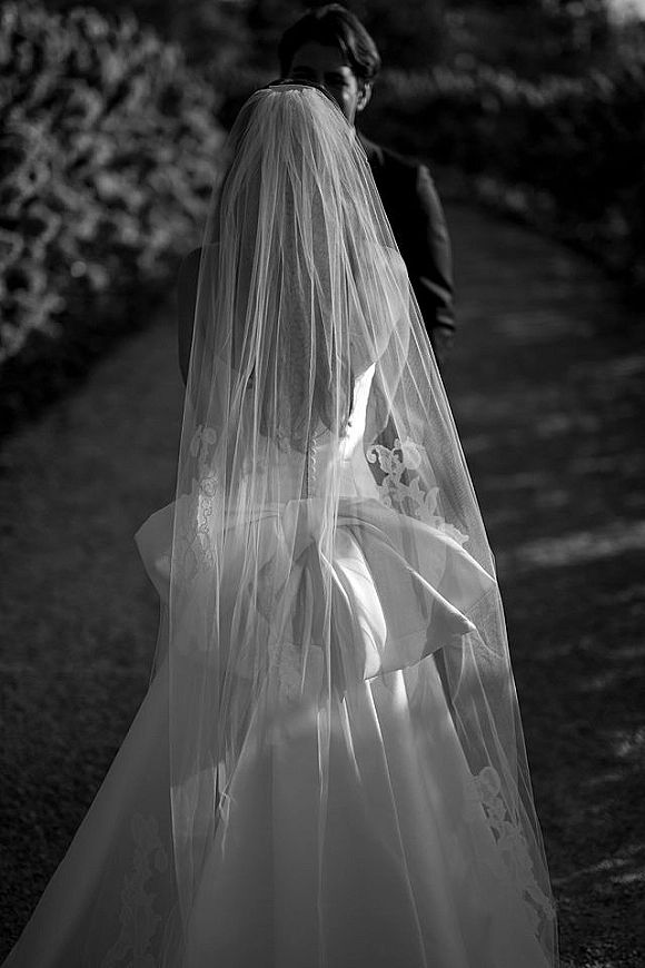 Wedding first look as bride from behind embraces groom on an outdoor tree-lined path, lace-trim cathedral veil flowing over her button-back gown