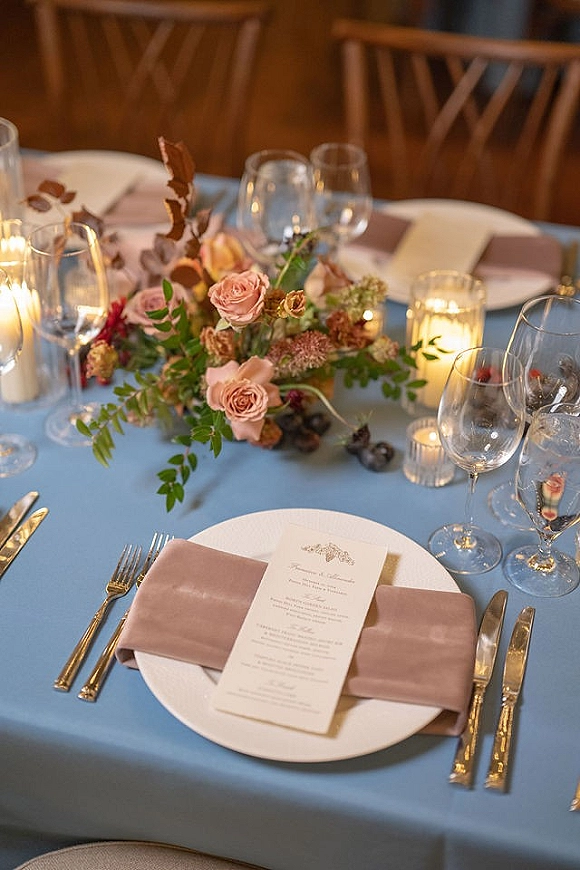 Reception tablescape with a wedding table centerpiece of roses and greenery, candlelight, gold flatware and blush napkins on blue linen, wooden chairs behind