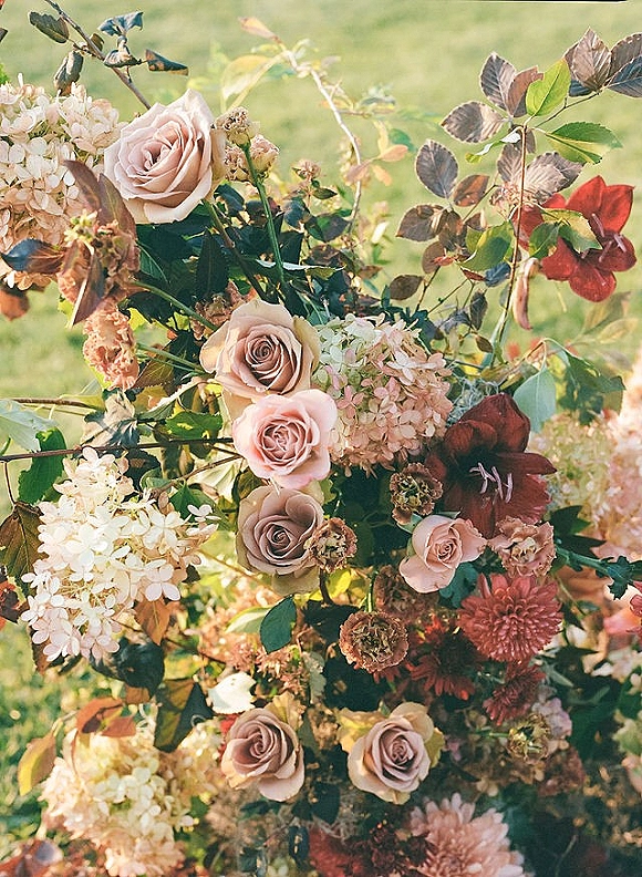 Wedding bouquet of blush roses and hydrangea with greenery and burgundy leaves, resting on sunlit grass with soft bokeh background