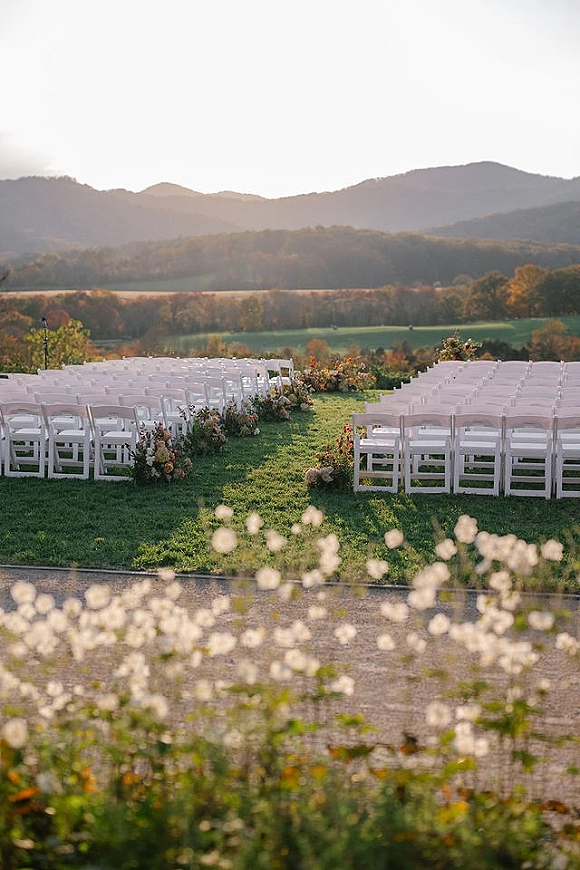 Outdoor ceremony setup with wedding ceremony chairs lining a floral-strewn aisle on a grass lawn overlooking mountains and valley