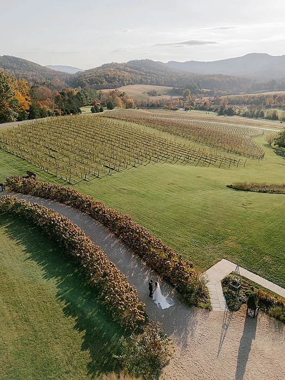 Couple portrait from above of bride and groom walking a gravel path through a vineyard, her veil trailing with mountains beyond