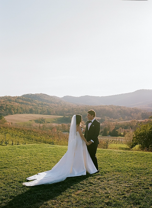 Couple portrait of bride and groom holding hands in a mountain wedding portrait, her long train and veil flowing over grass near vineyards